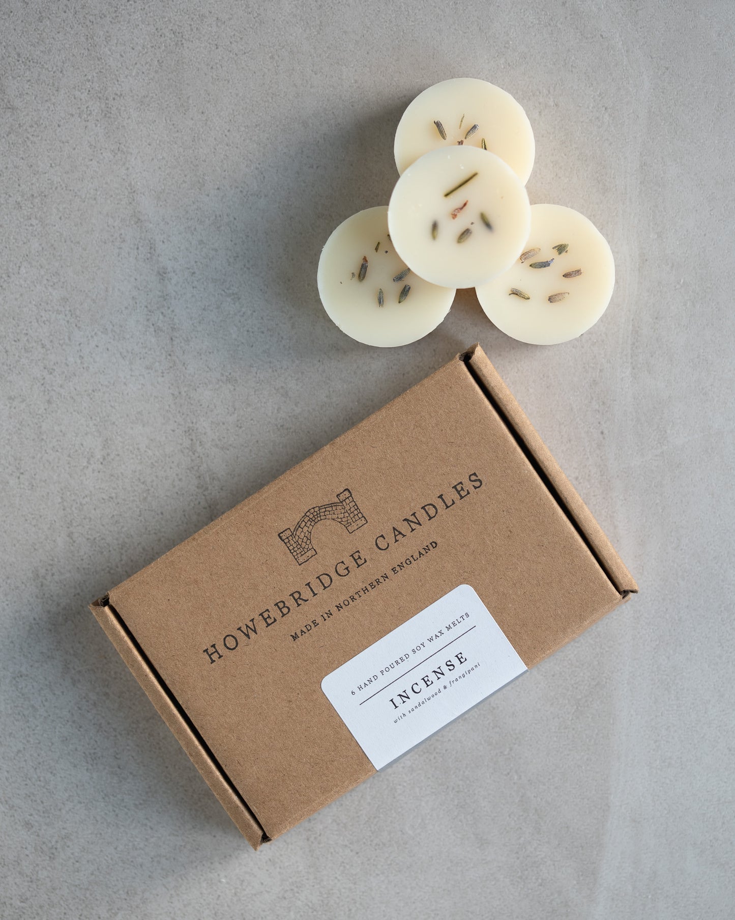 Howebridge Candles natural soy wax melts, stacked and sprinkled with dried lavender. Next to them is the wax melt packaging, a kraft box stamped with their logo and sealed with a scent descriptive label.