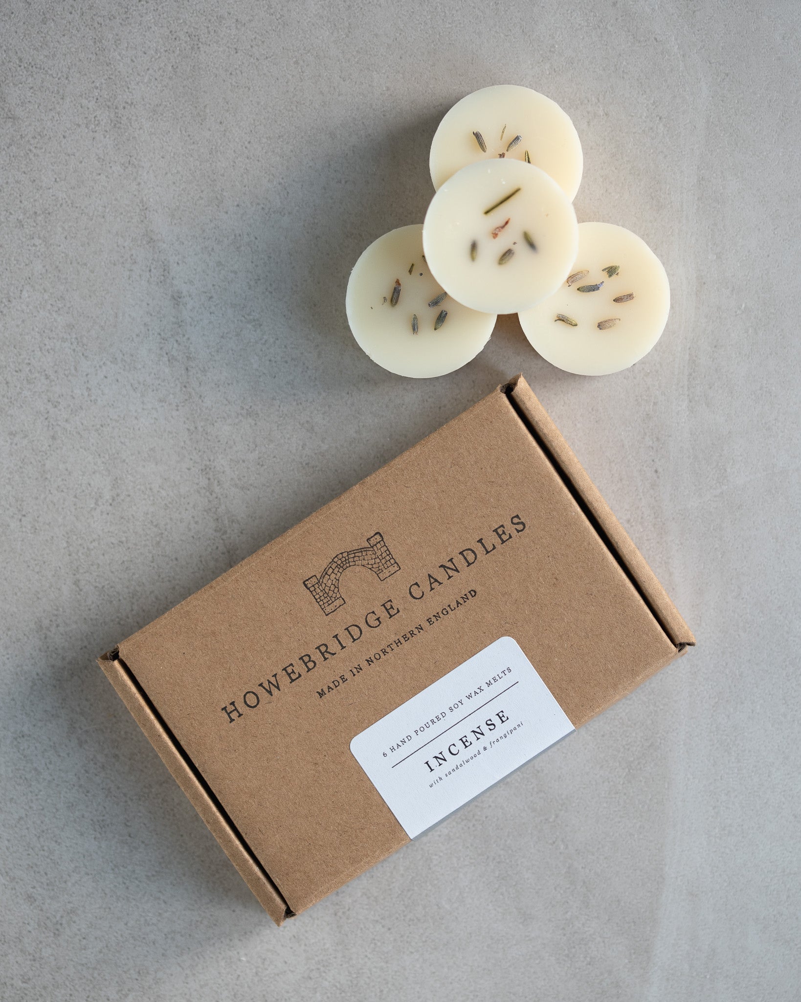 Howebridge Candles natural soy wax melts, stacked and sprinkled with dried lavender. Next to them is the wax melt packaging, a kraft box stamped with their logo and sealed with a scent descriptive label.