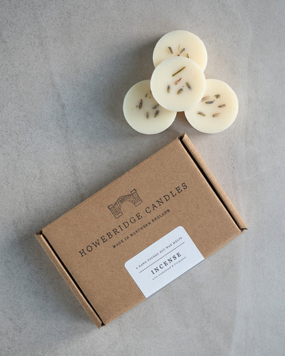Howebridge Candles natural soy wax melts, stacked and sprinkled with dried lavender. Next to them is the wax melt packaging, a kraft box stamped with their logo and sealed with a scent descriptive label.