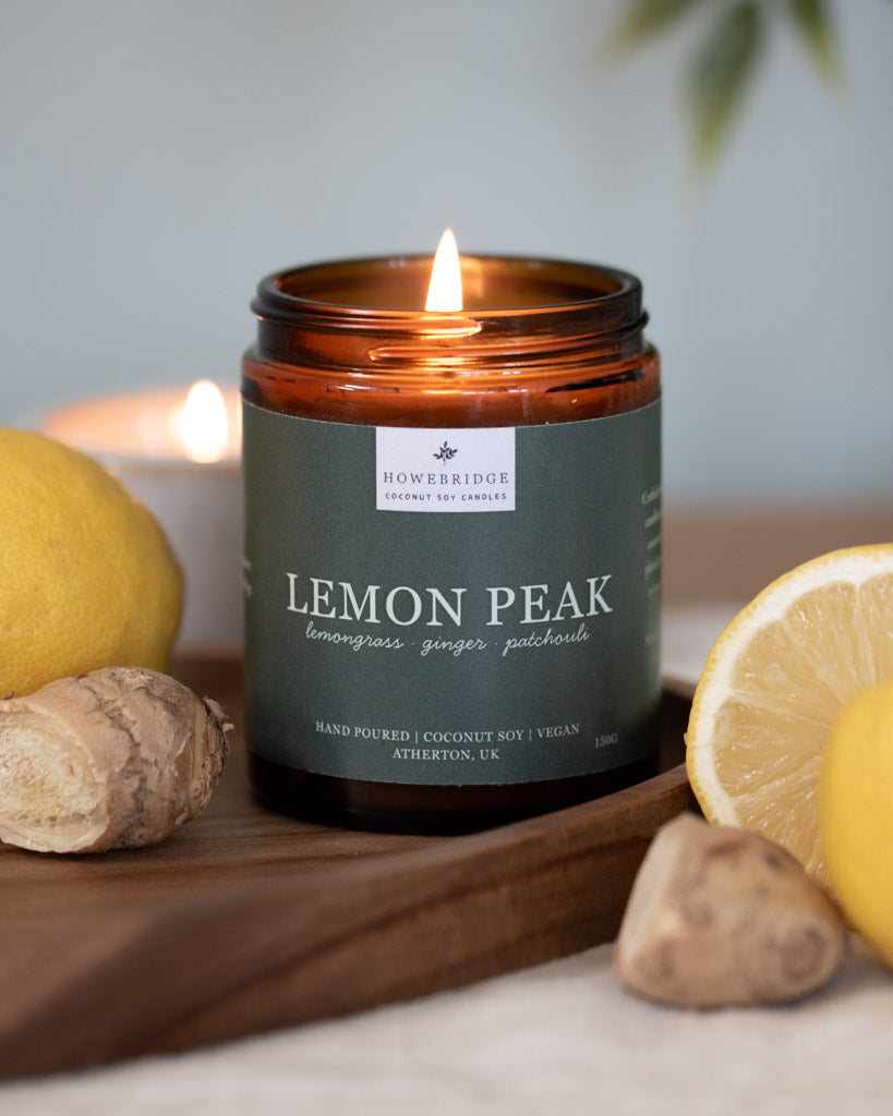 A lit Lemon Peak candle surrounded by lemons and ginger. A sage green wrap around label on a amber jar. Scent notes: lemongrass, ginger and patchouli. Hand poured, coconut soy and vegan. Poured in the Howe Bridge, North West, UK.