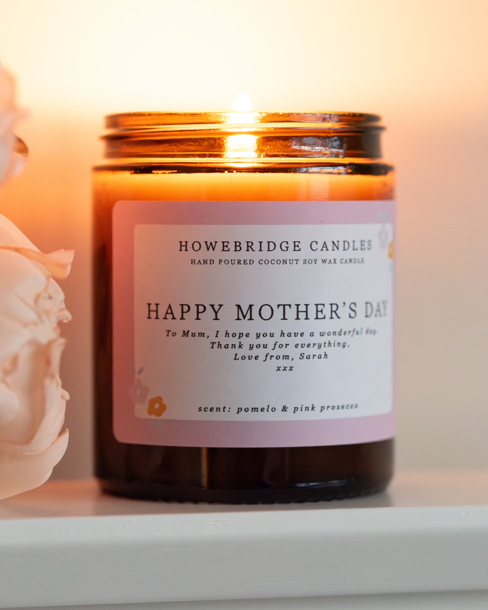 Personalised mother's day candle, made by Howebridge Candles. A glass amber jar  candle and label saying happy mothers day, lit and sat on a white shelf with pink flowers.
