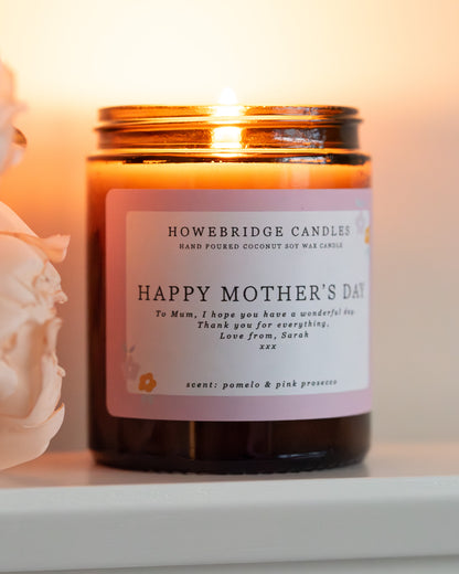 Personalised mother's day candle, made by Howebridge Candles. A glass amber jar  candle and label saying happy mothers day, lit and sat on a white shelf with pink flowers.