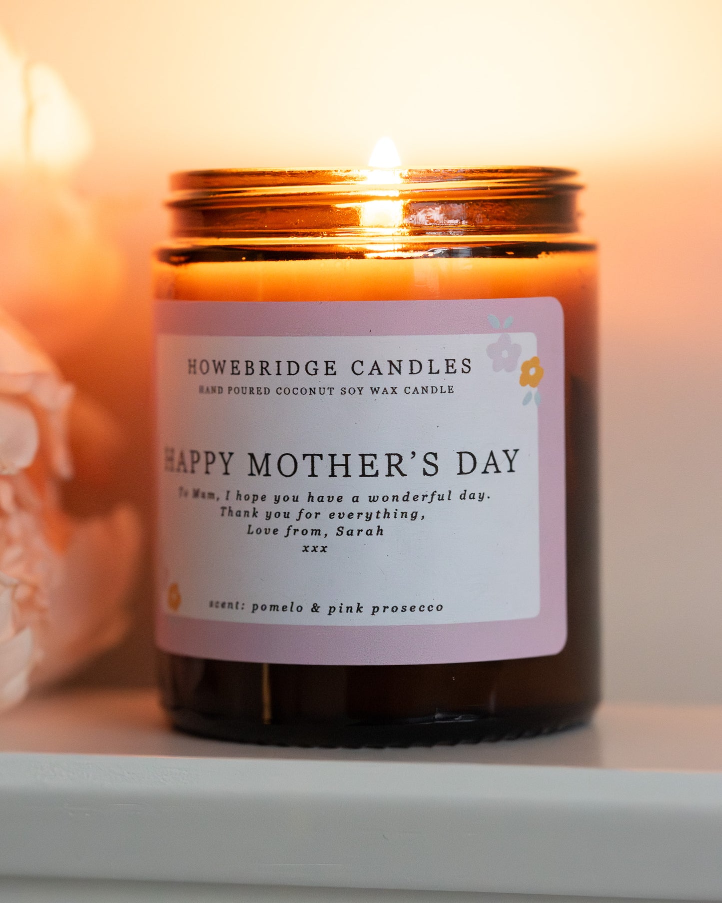 Personalised mother's day candle, made by Howebridge Candles. A glass amber jar  candle and label saying happy mothers day, lit and sat on a white shelf with pink flowers.