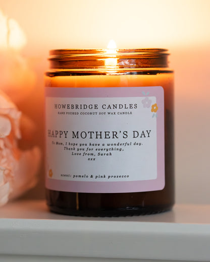 Personalised mother's day candle, made by Howebridge Candles. A glass amber jar  candle and label saying happy mothers day, lit and sat on a white shelf with pink flowers.
