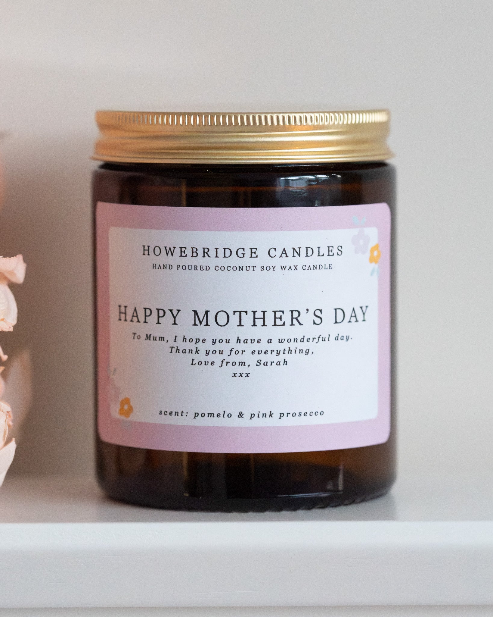 Personalised mother's day candle, made by Howebridge Candles. A glass amber jar  candle with gold lid and label saying happy mothers day, sat on a white shelf with pink flowers.