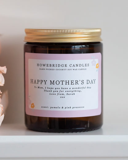 Personalised mother's day candle, made by Howebridge Candles. A glass amber jar  candle with gold lid and label saying happy mothers day, sat on a white shelf with pink flowers.