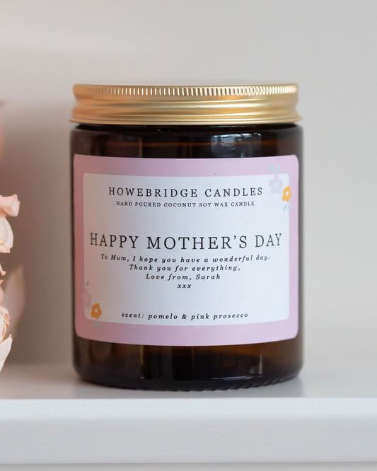 Personalised mother's day candle, made by Howebridge Candles. A glass amber jar  candle with gold lid and label saying happy mothers day, sat on a white shelf with pink flowers.