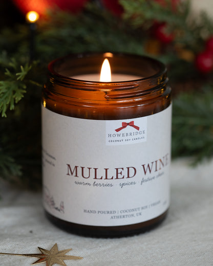 Mulled wine scented candle in a glass amber jar and gold lid, wrapped in a cream festive label featuring a red bow. Scent notes of warm berries, spices like cinnamon and festive cheer.