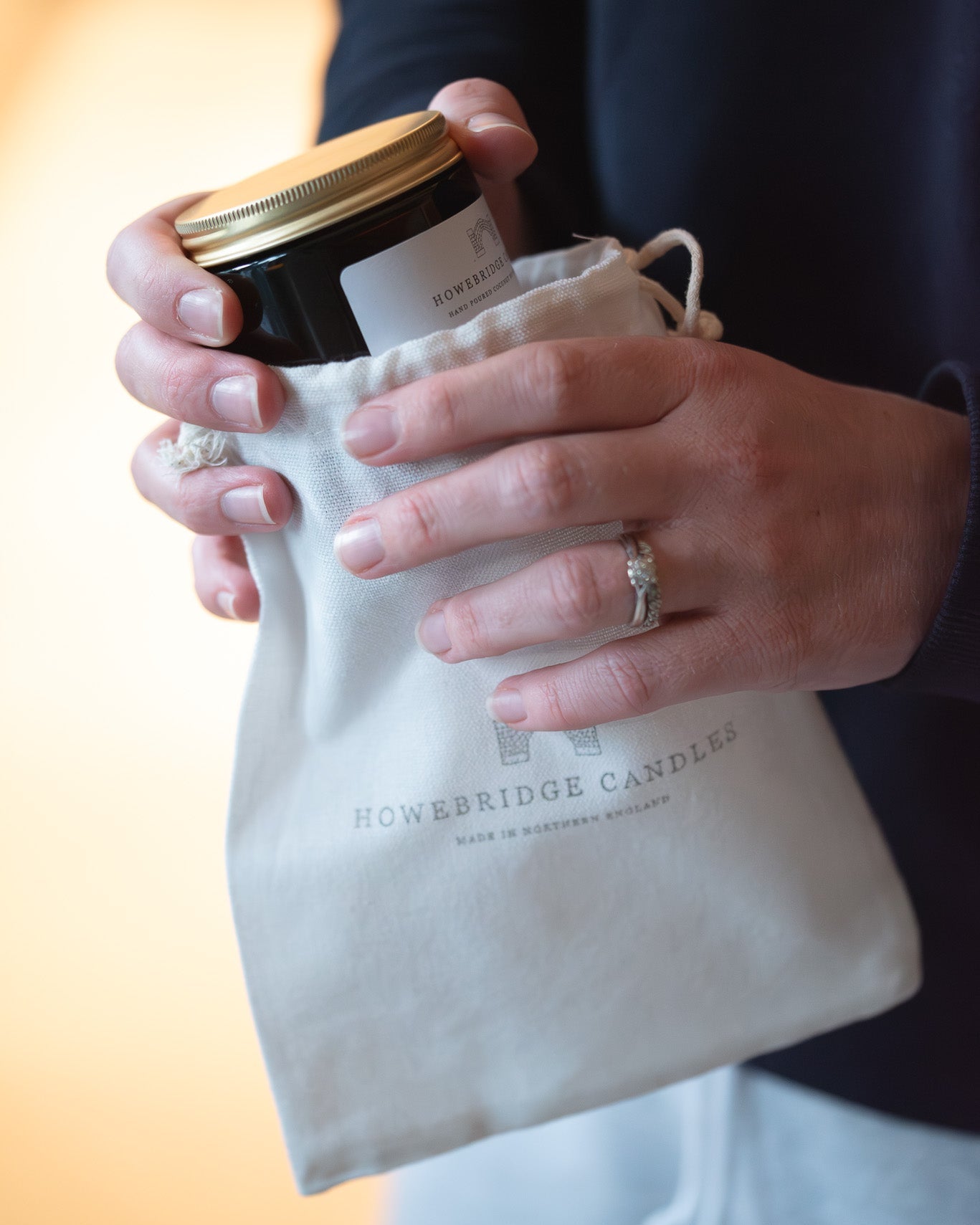 glass amber candle with gold lid, being pulled out of a cotton drawstring bag branded with the Howebridge Candles logo.