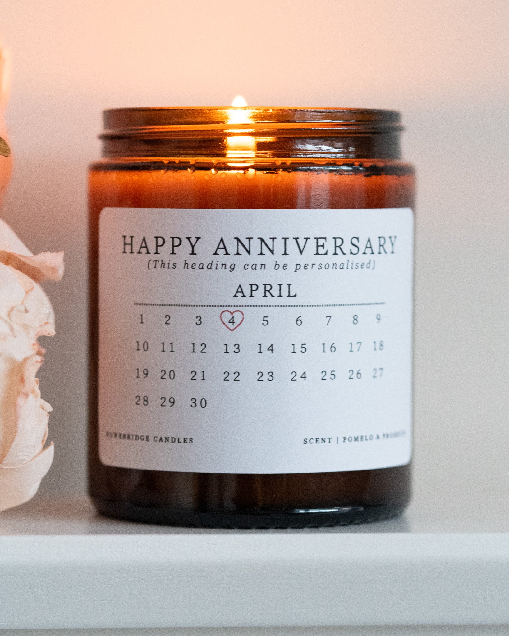 A personalised anniversary candle by Howebridge Candles. A glass amber jar and white label that features a happy anniversary heading and calendar. Lit and sat on a white shelf with pink flowers.