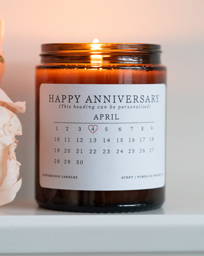 A personalised anniversary candle by Howebridge Candles. A glass amber jar and white label that features a happy anniversary heading and calendar. Lit and sat on a white shelf with pink flowers.