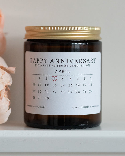 A personalised anniversary candle by Howebridge Candles. A glass amber jar with gold lid and white label that features a happy anniversary heading and calendar. Sat on a white shelf with pink flowers.