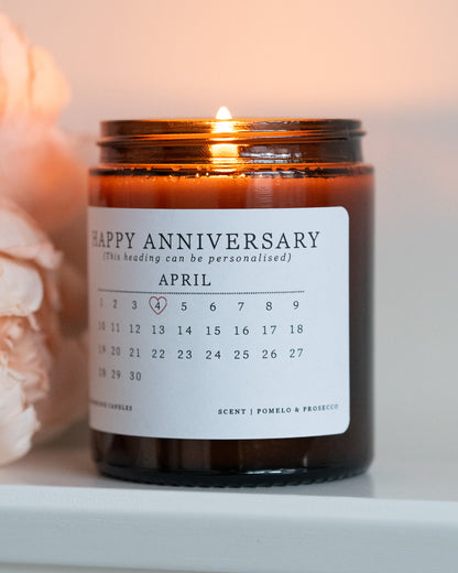 A personalised anniversary candle by Howebridge Candles. A glass amber jar and white label that features a happy anniversary heading and calendar. Lit and sat on a white shelf with pink flowers.