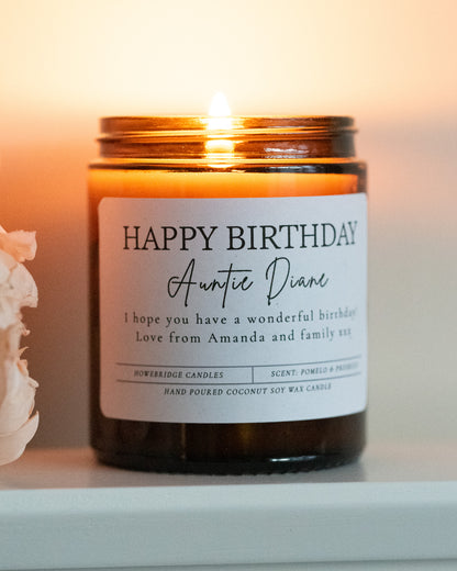 'Happy Birthday' Candle with name and message. Made by Howebridge Candles in a glass amber jar, it's lit and sat on a white shelf with pink flowers.
