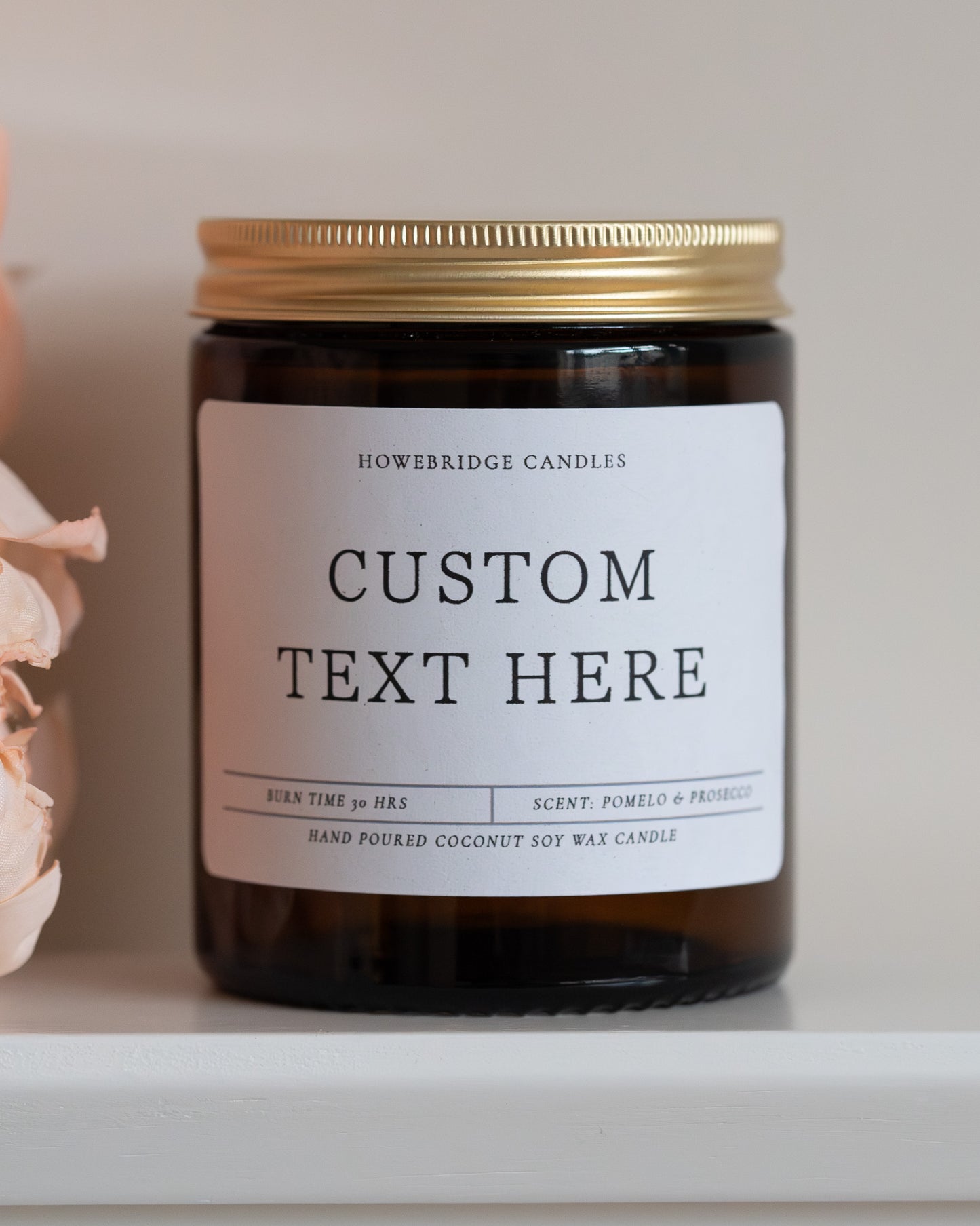 A amber jar with gold lid candle by Howebridge Candles. A white label that says 'custom text here' and sat on a white shelf with pink flowers.