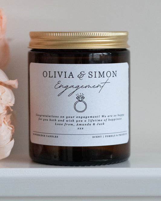 A personalised engagement candle gift by Howebridge Candles, featuring a glass amber jar, gold lid and white label that has names, a ring and a personalised message. Sat on a white shelf with pink flowers.