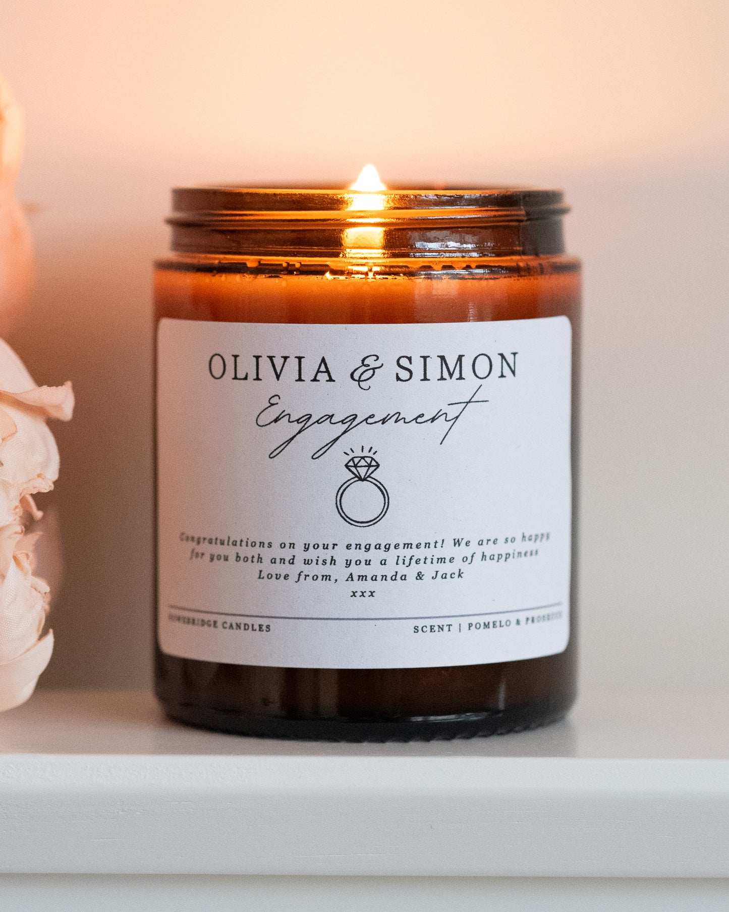 A personalised engagement candle gift by Howebridge Candles, featuring a glass amber jar, gold lid and white label that has names, a ring and a personalised message. Lit and sat on a white shelf with pink flowers.