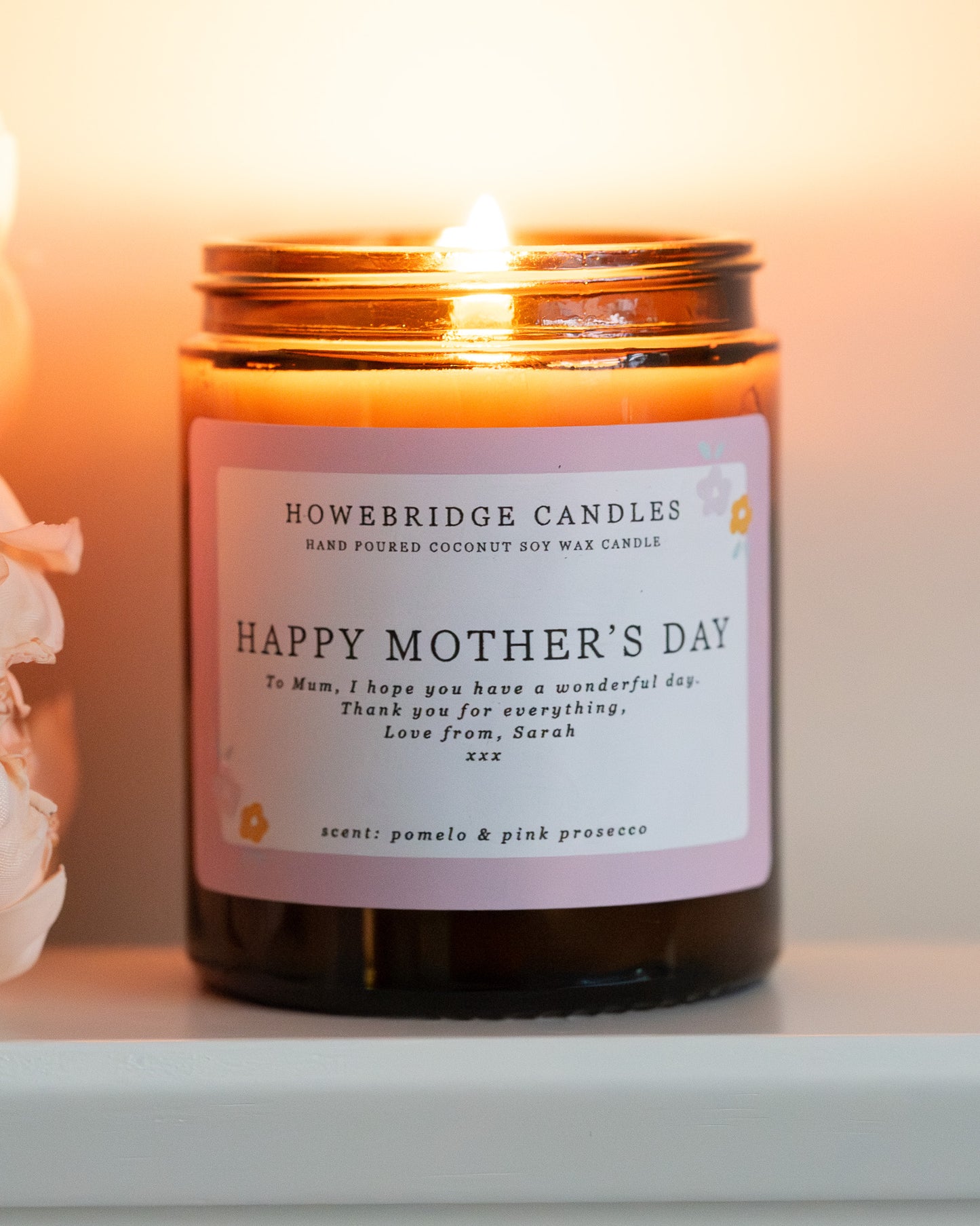 Personalised mother's day candle, made by Howebridge Candles. A glass amber jar  candle and label saying happy mothers day, lit and sat on a white shelf with pink flowers.