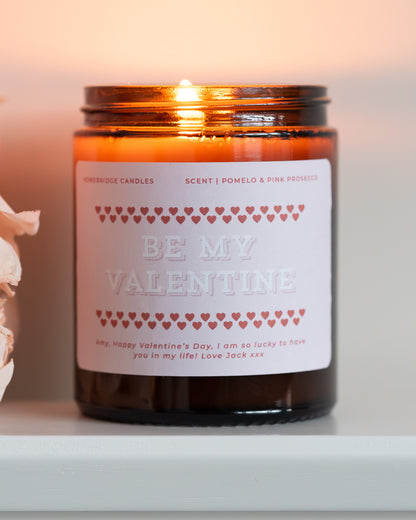 Glass Amber candle jar with pink 'Be My Valentine' label, lit and on a white surface, with some pink flowers.