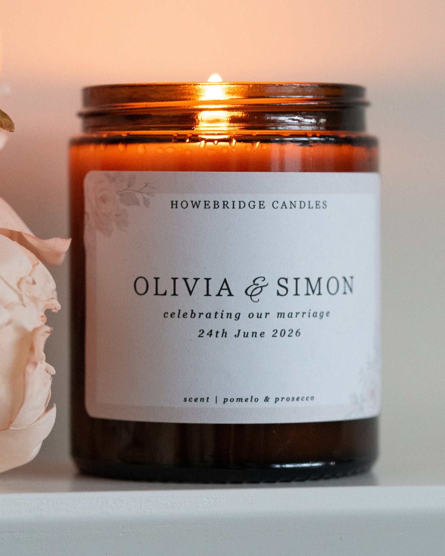 A wedding day candle by Howebridge Candles. A glass amber jar candle featuring a floral label. Lit and sat on a white shelf with pink flowers.