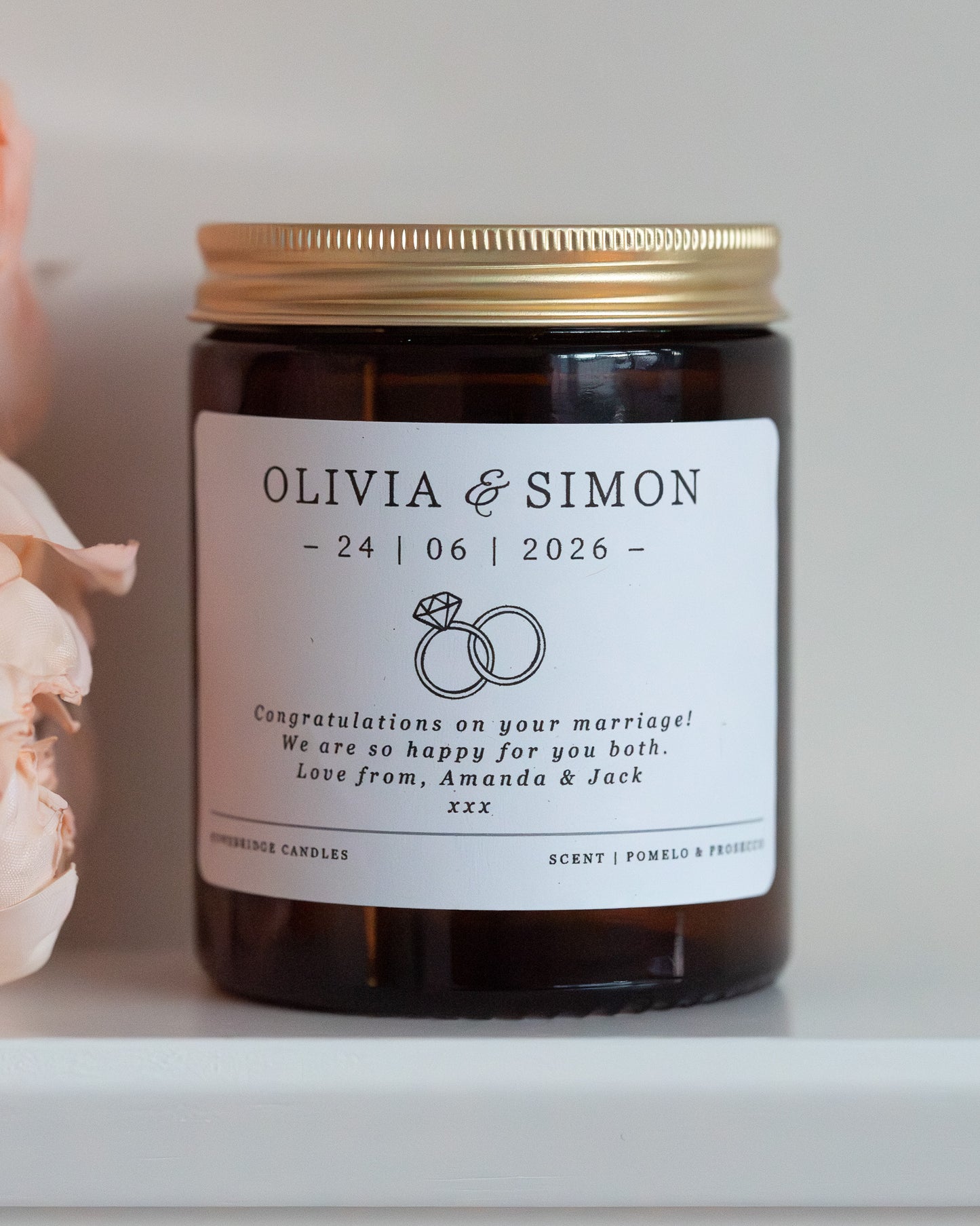 A personalised candle for a wedding day gift. Made by Howebridge Candles in a glass amber jar and gold lid, featuring a white label. Sat on a white shelf with pink flowers.