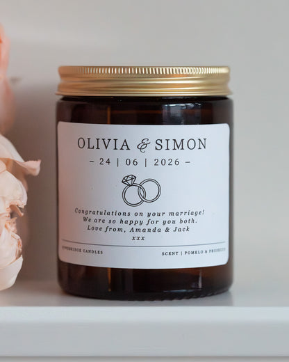 A personalised candle for a wedding day gift. Made by Howebridge Candles in a glass amber jar and gold lid, featuring a white label. Sat on a white shelf with pink flowers.