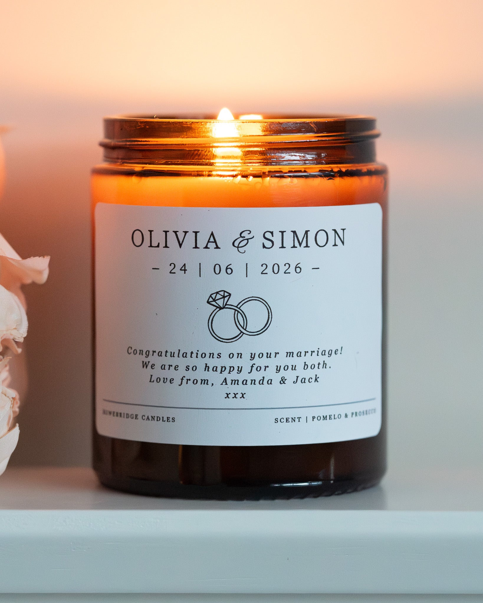 A personalised candle for a wedding day gift. Made by Howebridge Candles in a glass amber jar featuring a white label. Lit and sat on a white shelf with pink flowers.