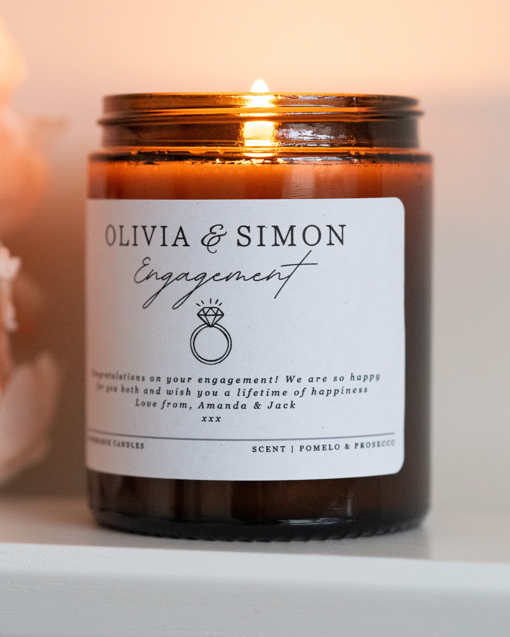 A personalised engagement candle gift by Howebridge Candles, featuring a glass amber jar, gold lid and white label that has names, a ring and a personalised message. Lit and sat on a white shelf with pink flowers.