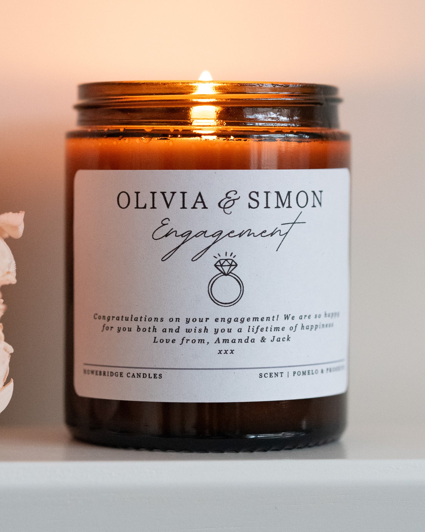A personalised engagement candle gift by Howebridge Candles, featuring a glass amber jar, gold lid and white label that has names, a ring and a personalised message. Lit and sat on a white shelf with pink flowers.