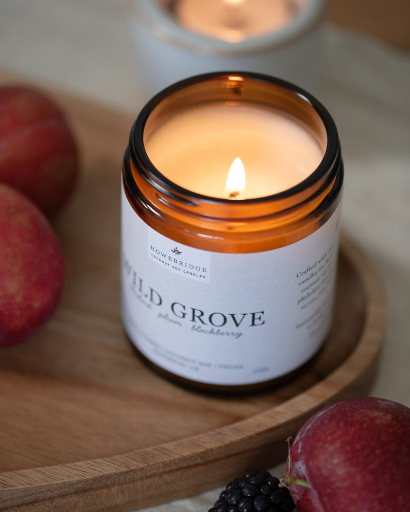 Wild Grove candle lit on a wooden board, surrounded by summer fruits. In a glass amber jar with a off white natural label. Scent: Rhubarb, plum and blackberries. Our bestseller all year round.