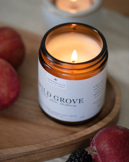 Wild Grove candle lit on a wooden board, surrounded by summer fruits. In a glass amber jar with a off white natural label. Scent: Rhubarb, plum and blackberries. Our bestseller all year round.