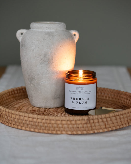 Rhubarb and plum scented candles by Howebridge candles, sitting on a rattan tray with a vase, it's lid and a match stick. The candle is lit in a glass amber jar.