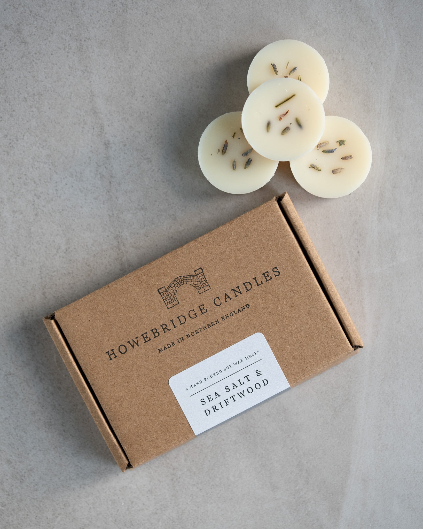 Howebridge Candles sea salt & driftwood soy wax melts, stacked and sprinkled with dried lavender. Next to them is the wax melt packaging, a kraft box stamped with their logo and sealed with a scent descriptive label.