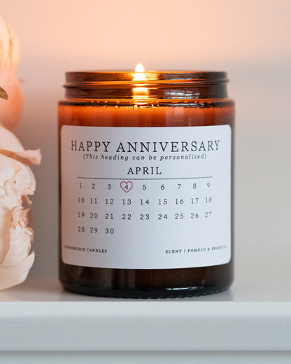 A personalised anniversary candle by Howebridge Candles. A glass amber jar and white label that features a happy anniversary heading and calendar. Lit and sat on a white shelf with pink flowers.