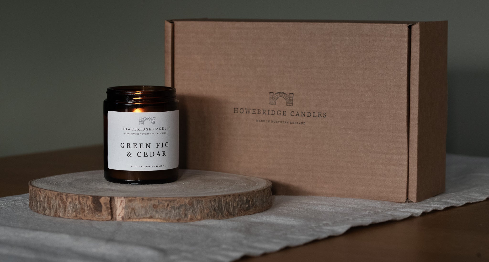 Candle labeled 'Green Fig & Cedar' by Howebridge Candles on a wooden coaster with a matching box in the background.