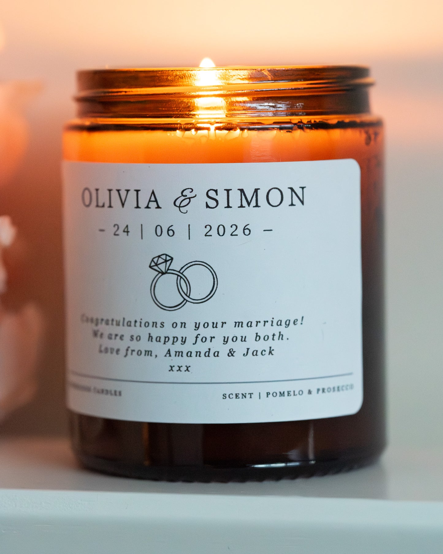 A personalised candle for a wedding day gift. Made by Howebridge Candles in a glass amber jar featuring a white label. Lit and sat on a white shelf with pink flowers.