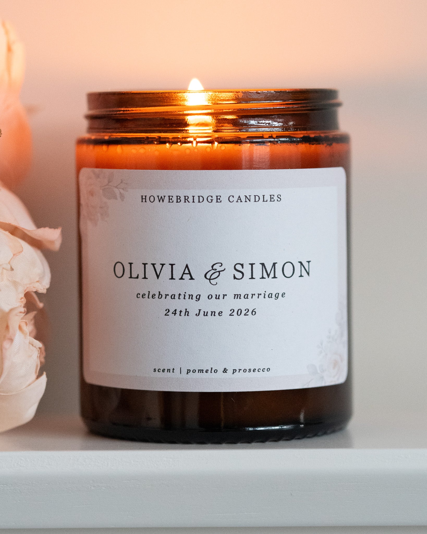 A wedding day candle by Howebridge Candles. A glass amber jar candle featuring a floral label. Lit and sat on a white shelf with pink flowers.