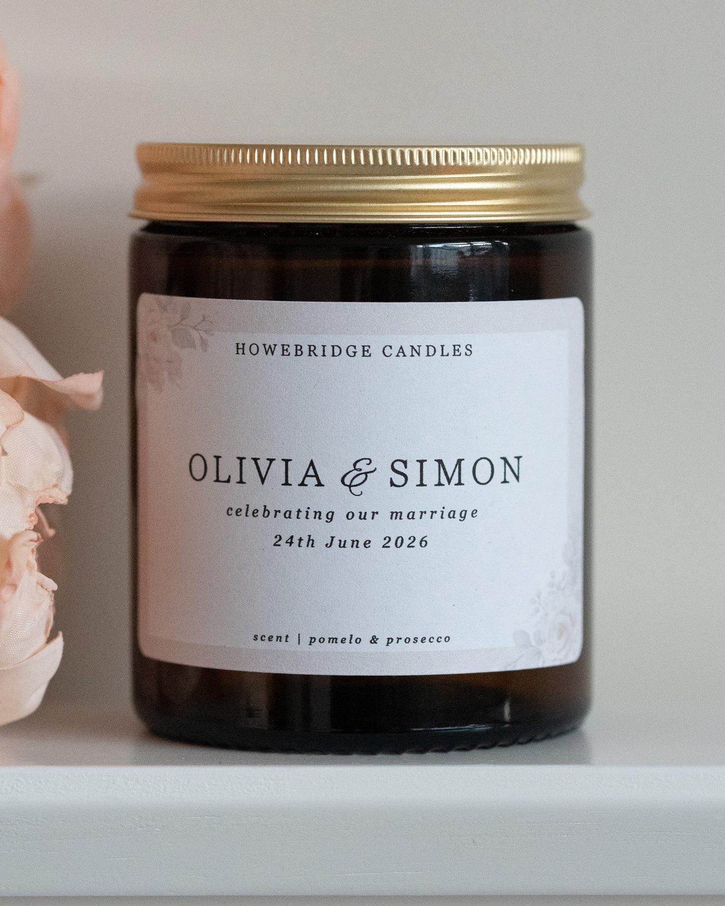 A wedding day candle by Howebridge Candles. A glass amber jar and gold lid featuring a floral label. Sat on a white shelf with pink flowers.
