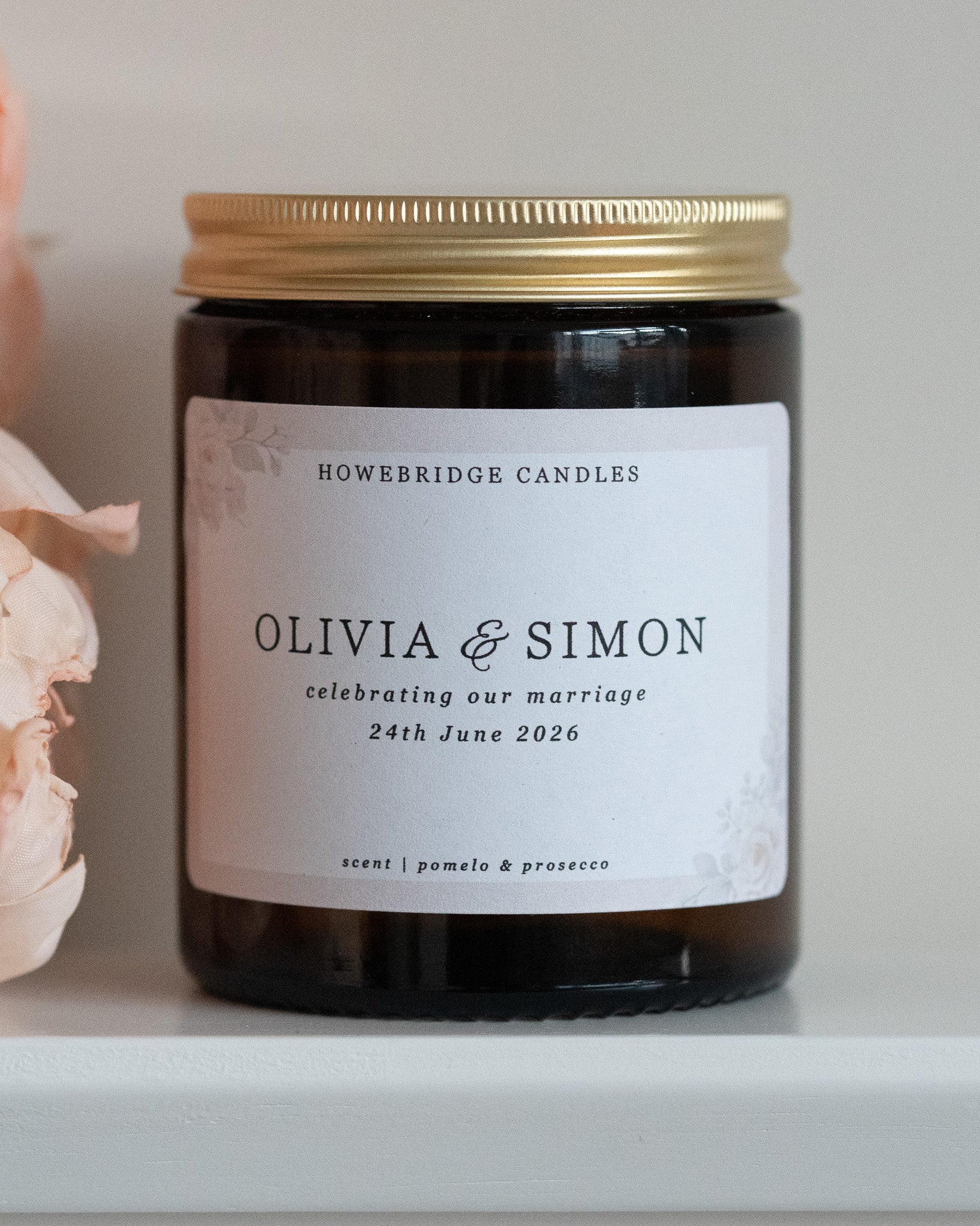 A wedding day candle by Howebridge Candles. A glass amber jar and gold lid featuring a floral label. Sat on a white shelf with pink flowers.
