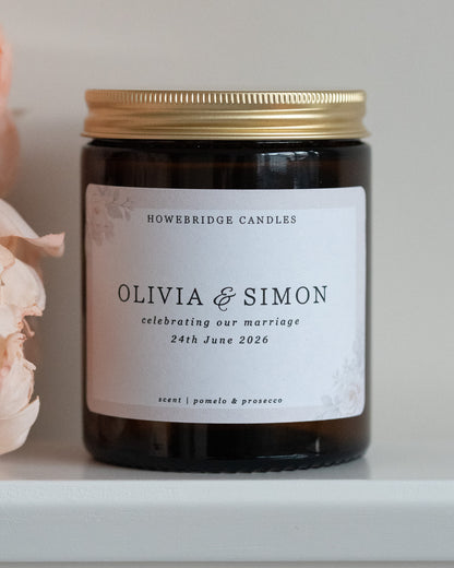 A wedding day candle by Howebridge Candles. A glass amber jar and gold lid featuring a floral label. Sat on a white shelf with pink flowers.