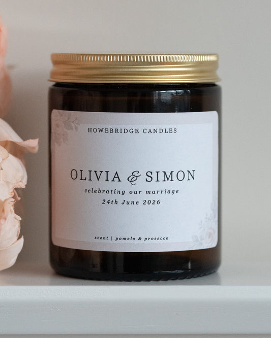 A wedding day candle by Howebridge Candles. A glass amber jar and gold lid featuring a floral label. Sat on a white shelf with pink flowers.